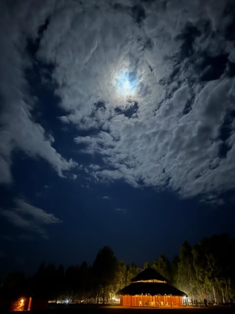 A full moon through clouds above a lit thatched pavilion.